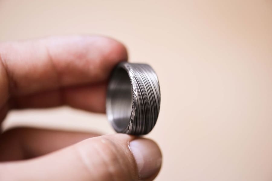 Damascus steel ring (jewellery) held out by fingertips with a pale beige background.