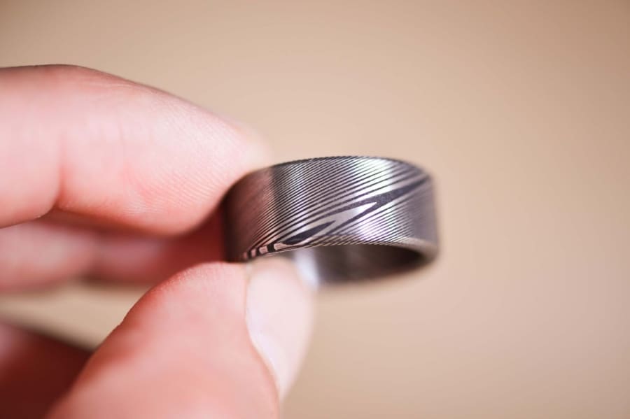 Damascus steel ring (jewellery) held out by fingertips with a pale beige background.