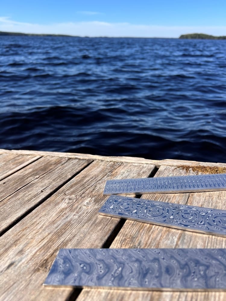 Three polished and patterned bars of Damascus steel are lying on a weathered wooden dock with dark water in the background.