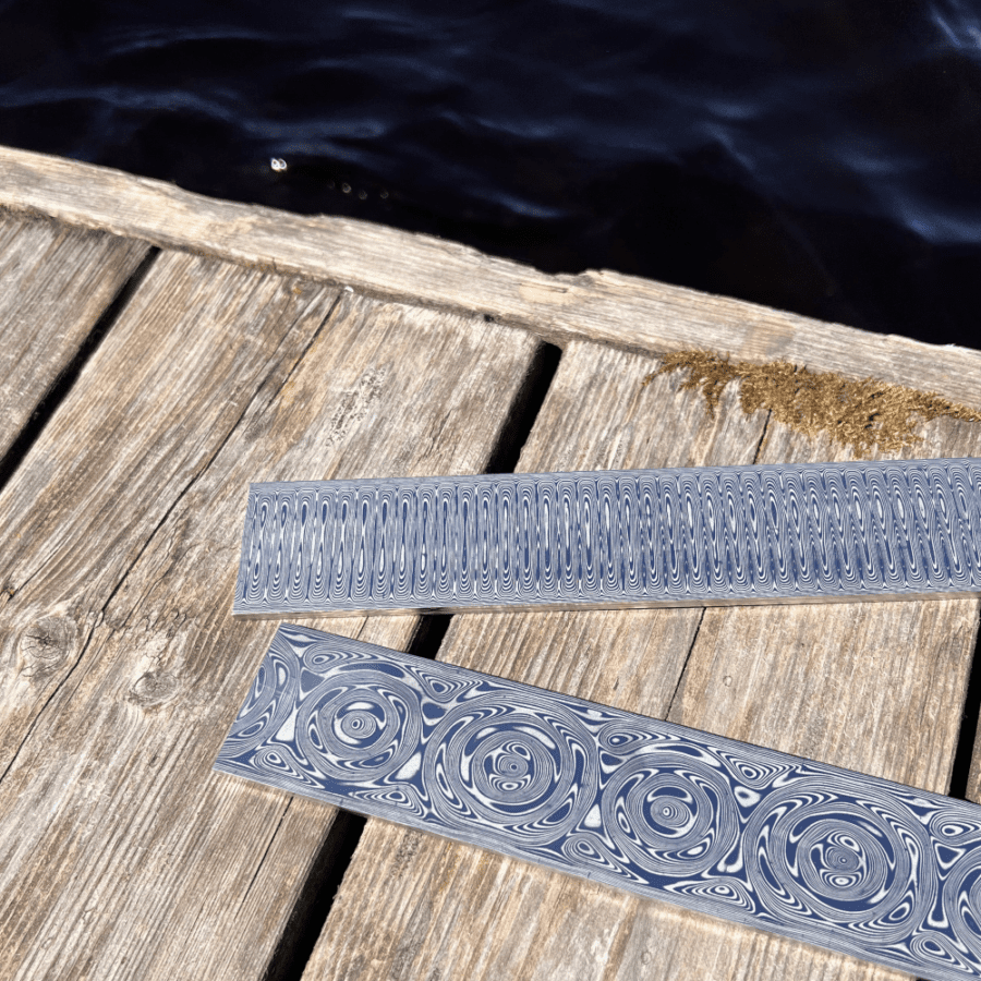 Two polished and patterned bars of Damascus steel are laying on a weathered wooden dock with dark water in the background.