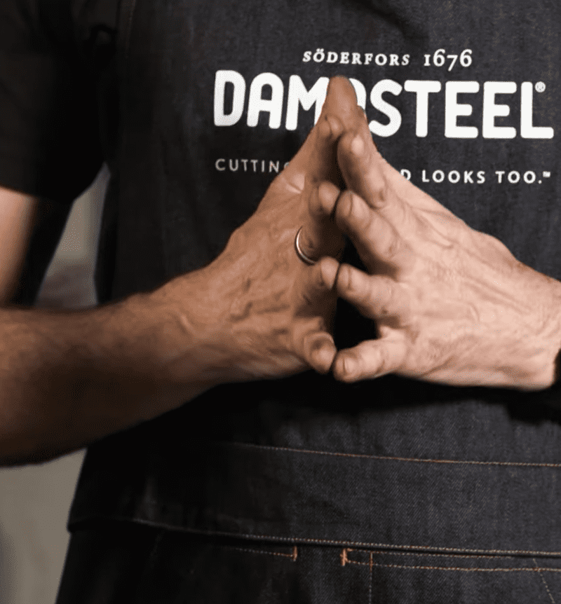 Hands of one man touching in front of a denim apron with log of Damasteel